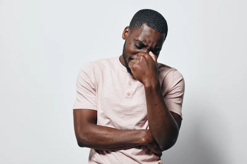 African man with dark skin wearing light pink shirt standing against white background with eyes closed and hand covering face expressing stress or sadness in studio.
