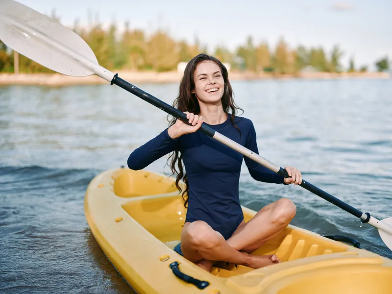 Young woman paddling a yellow kayak on calm water, smiling as she grips the paddle, wearing a navy suit, sunlight glints, creating a serene outdoor activity.