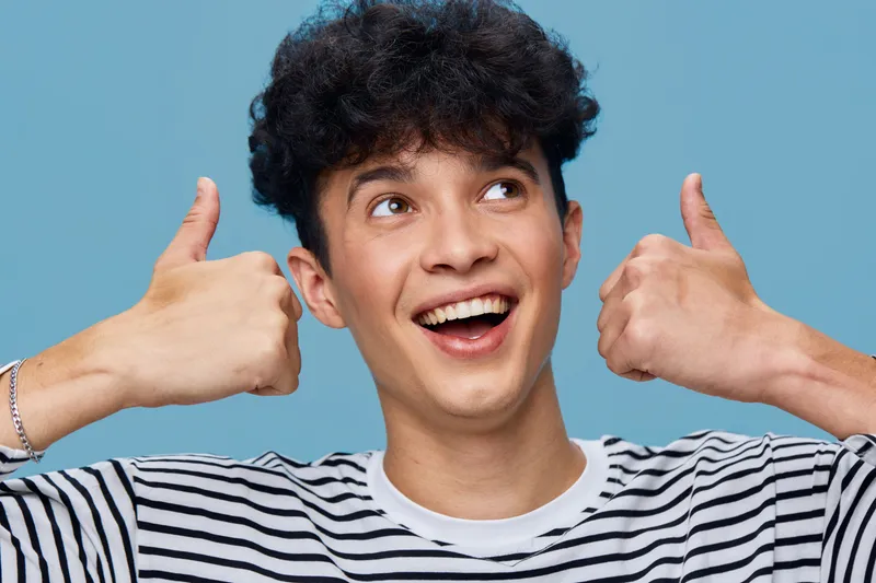 smiling man showing thumbs up wearing striped shirt against blue background, cheerful young adult with curly hair expressing positivity and happiness