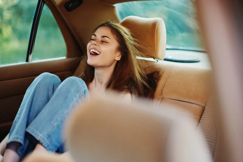 Joyful young woman laughing in a car, wearing casual clothing, embodying carefree happiness with a vibrant outdoor background, perfect for travel or lifestyle themes.