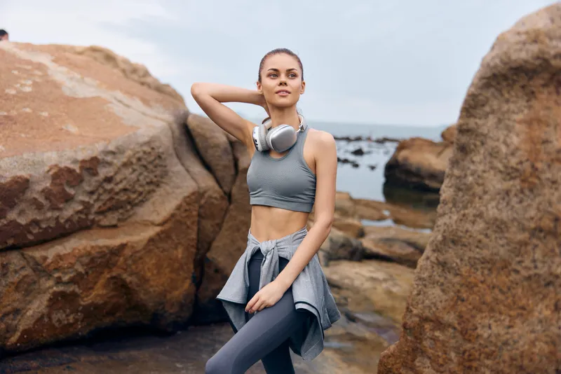 Fit young woman in athletic wear poses confidently by the rocky beach, showcasing a healthy lifestyle and outdoor fitness concept with headphones