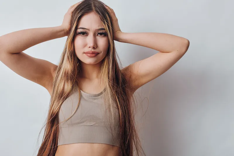 Young woman with long blonde hair wearing a sleeveless crop top posing with arms raised behind head against plain background, casual and confident expression for portrait.