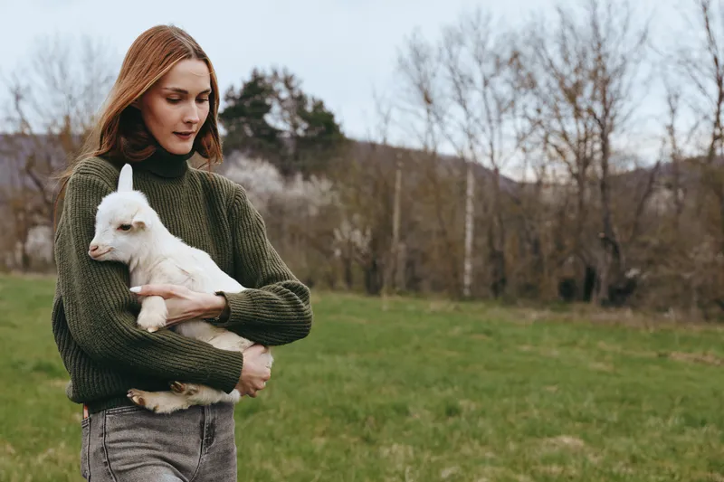 Young woman in a green knit sweater holds a fluffy white goat kid in an open field, expressing tender companionship, rustic lifestyle portrait, natural light
