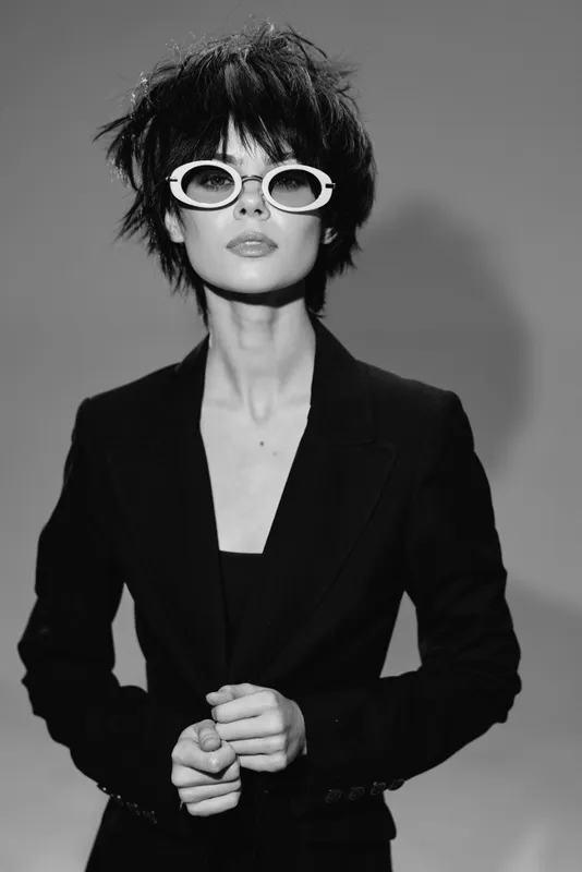 Stylish woman in black suit with white sunglasses striking confident pose in black and white portrait.