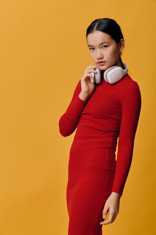 Woman in red dress with white headphones posing, looking at camera against yellow background, fashion portrait with modern style and casual elegance in studio lighting.