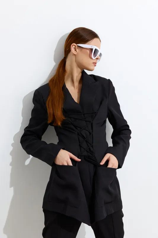 Stylish woman wearing oversized black blazer and white sunglasses posing confidently against white background in minimalistic fashion portrait with natural lighting.