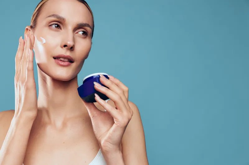 A young woman with light skin applies white cream to her cheek, holding a blue jar, as a soft blue background creates a calm skincare mood.