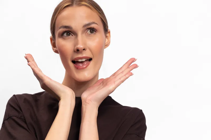 Young woman with blonde hair and expressive face looks surprised with hands near chin on white background. Concept of emotion, reaction, and positive expression.