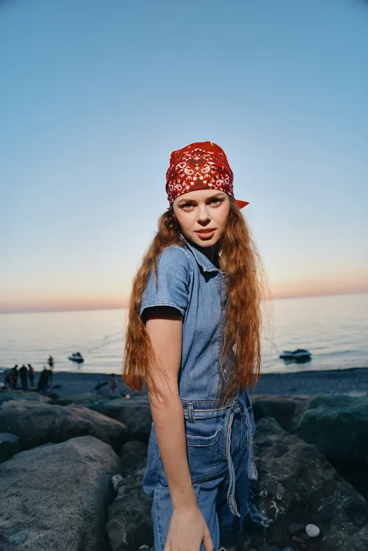 Lifestyle woman in modern boho-western denim street style stands by the ocean at sunset with a red bandana, featuring film color tones and relaxed confident expression.