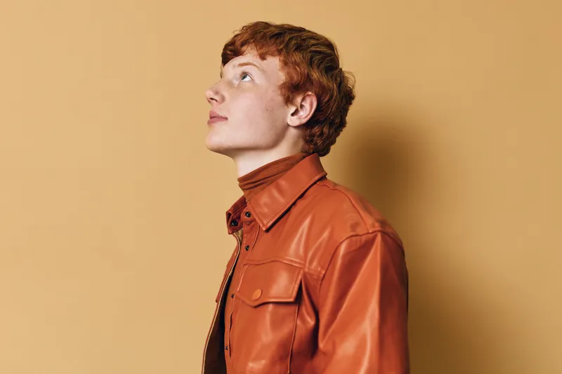 Young man with red hair wearing brown leather jacket looking upward on beige background. Profile portrait with thoughtful expression and trendy casual style in studio light.