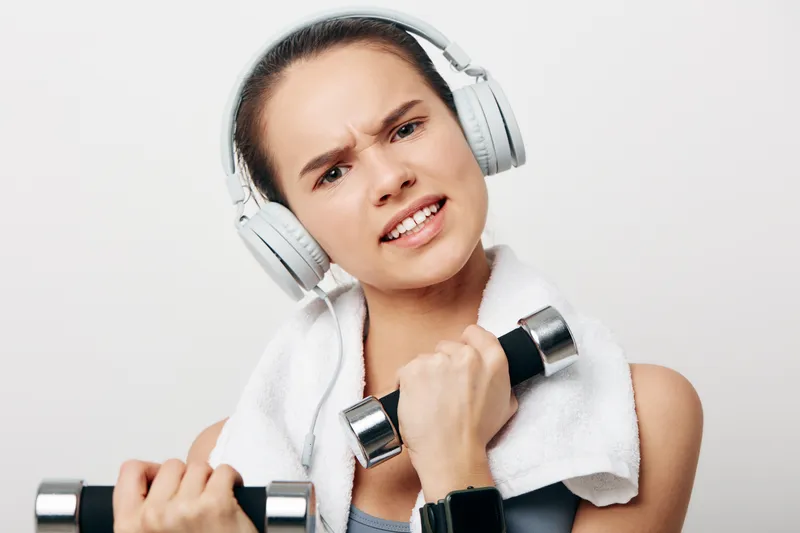 Portrait of a young woman fitness enthusiast wearing headphones and a towel, gripping chrome dumbbells with an intense focused expression against a clean white studio background.