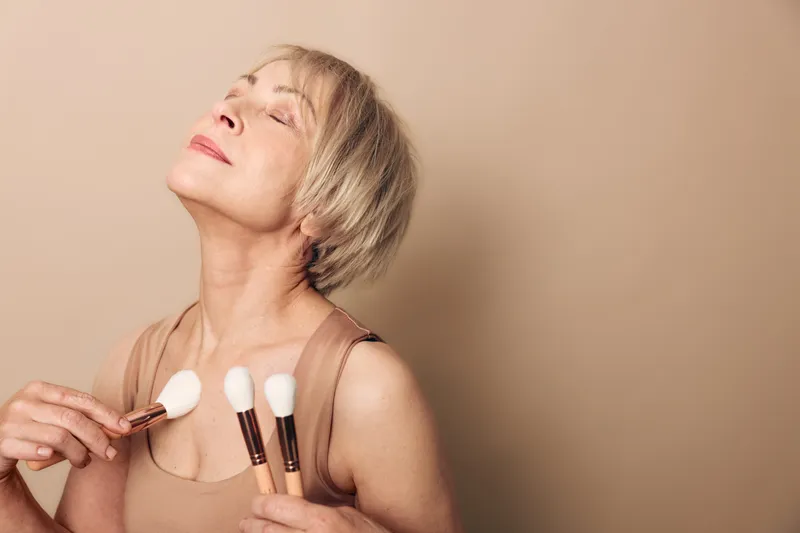 Mature woman with short blonde hair holds makeup brushes near chest, eyes closed in relaxed pose on beige background. Concept of beauty routine, self-care, and confidence in aging skin.