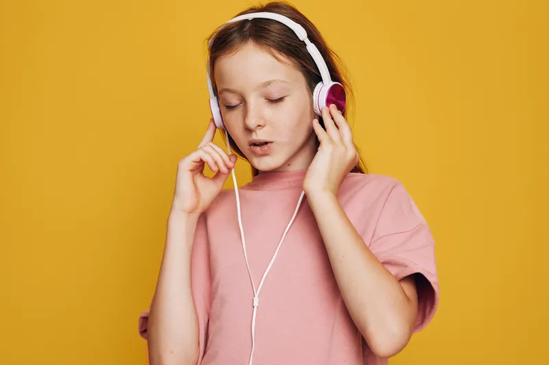 Young girl wearing pink t-shirt and white headphones listens to music with eyes closed against yellow background. Studio portrait capturing calm and focus. Concept of music and relaxation.