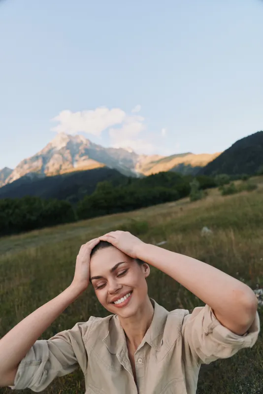 Meadow scene with a smiling woman in a beige shirt, hands on her head, sunlit grass swaying, conveying a relaxed outdoor lifestyle