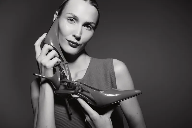 Blonde caucasian woman with light skin holds shiny high heel shoes near face in studio on dark background.
