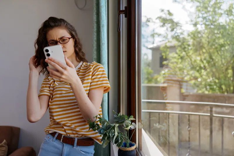Smiling woman in a striped shirt using smartphone by an open window at home, showcasing casual lifestyle and selfcare moment, natural light highlighting a relaxed mood and warmth throughout space