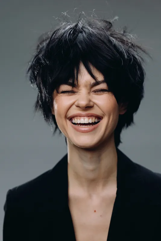 Smiling woman with short black hair laughing joyfully in studio with gray background wearing black jacket