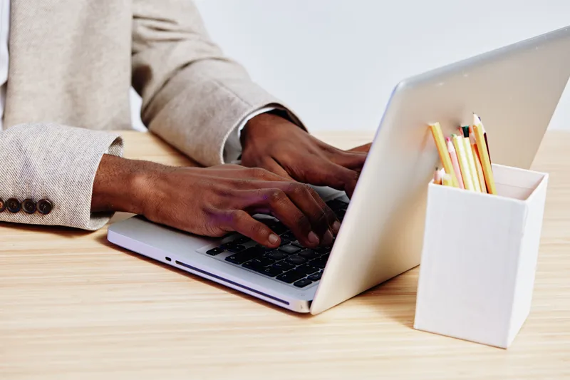Professional man using laptop at workspace on wooden desk, with white container holding colored pencils, dressed in beige blazer, close-up of hands, modern office environment, work and creativity conc