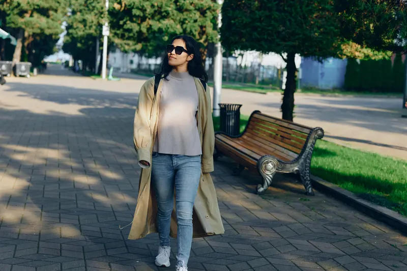 Woman in a long beige coat and sunglasses walks along a park path, casual street fashion in an outdoor setting with benches, trees, and soft sunlight.