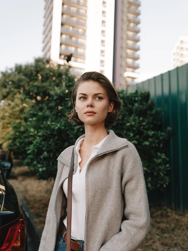A calm woman stands in an urban setting with tall buildings in the background. She wears a light coat and casual blouse, looking ahead with focus and confidence, creating a poised and contemporary