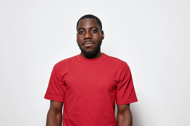 African American man with dark skin wearing red t shirt standing against white background looking at camera with neutral expression and short hair in studio portrait.