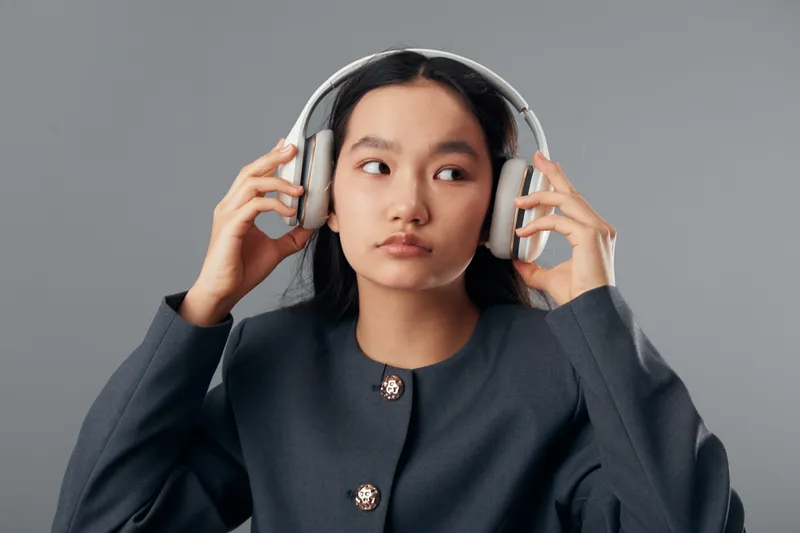 Young woman wearing headphones, listening to music or audio content with thoughtful expression on gray background. Casual style, technology, and entertainment concept in portrait.