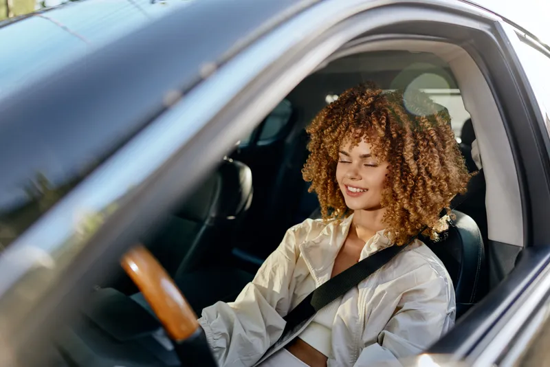 Smiling woman with curly hair sits in a car wearing a beige jacket and seatbelt, enjoying a sunny day with relaxed and happy expression inside modern vehicle.