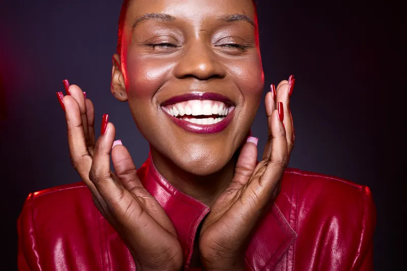 Close-up of happy black woman with dark skin smiling with closed eyes, wearing red leather jacket and matching lipstick. Concept of joy, beauty, fashion, and confident female model.