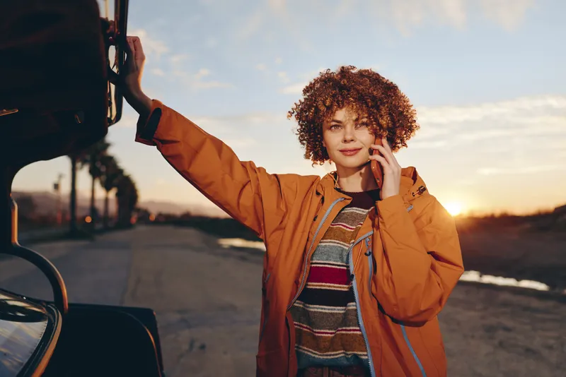 Woman outdoors lifestyle at sunset, curly haired woman smiles near car, wearing orange jacket, adjusting hair by ear, warm light, roadside background, casual travel mood evening breeze