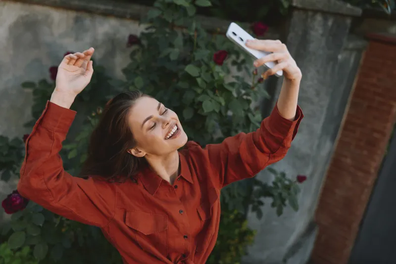 Young woman in a red shirt takes a selfie outdoors with a joyful smile and raised arms