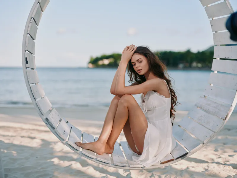 A young woman sits in a white circular beach swing by the shore, barefoot, wearing a light dress, looking down with a peaceful, contemplative expression.