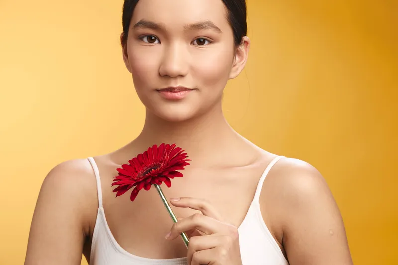 asian woman holds a flower in a studio setting. she gazes softly at the camera, radiating calm elegance and gentle charm. warm light highlights smooth skin and delicate petals, creating a serene