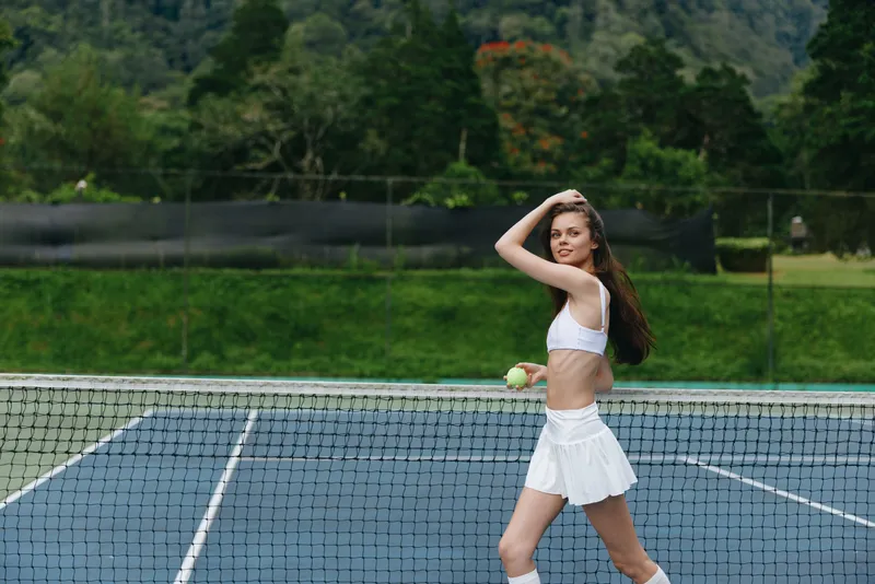 Young athletic woman in white sportswear joyfully playing tennis on outdoor court, embodying energy and health through sport and fitness