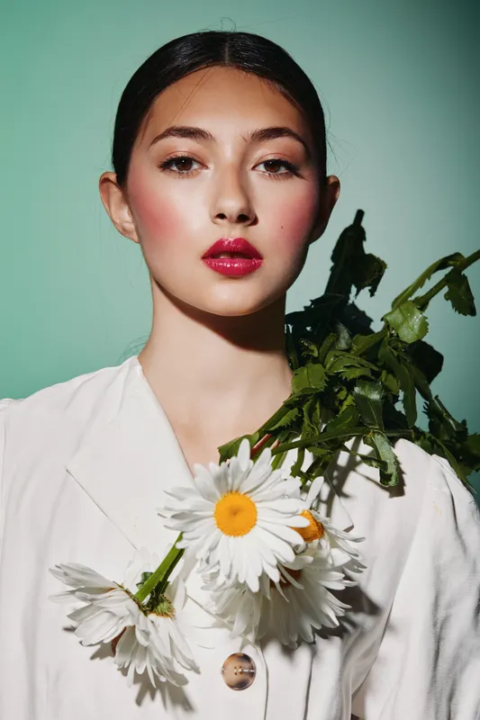 Elegant young woman with bright red lips and minimal makeup holds a bouquet of white daisies on her shoulder. Clean background highlights creative fashion and beauty concept.