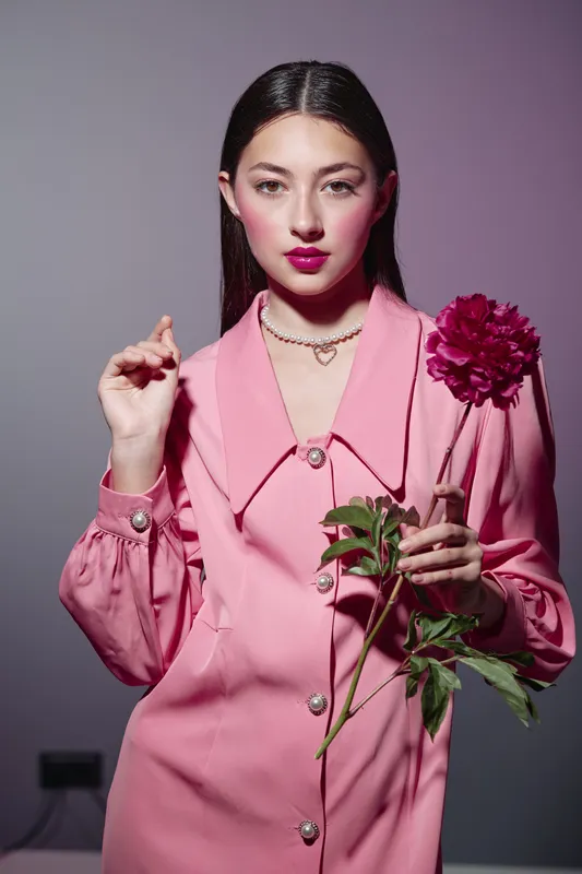 Young woman in pink vintage coat holding dark pink flower. Bold lip makeup and stylish pearl necklace create a strong fashion and beauty photo concept in studio light.