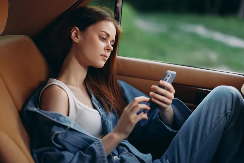 Young woman in denim outfit using smartphone in car, relaxed and casual, green nature background, modern lifestyle concept.