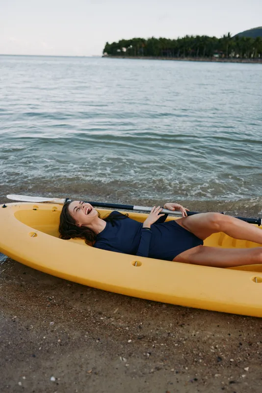 A relaxed woman lies on a bright yellow kayak along a pebbled shore, wearing a navy swimsuit, enjoying warm sun, gentle waves, and tranquil seaside scenery.