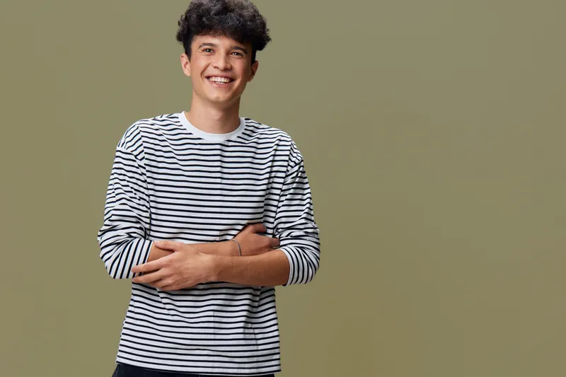 smiling man wearing striped shirt stands against beige background with arms crossed, youthful expression, casual style and relaxed mood captured vividly