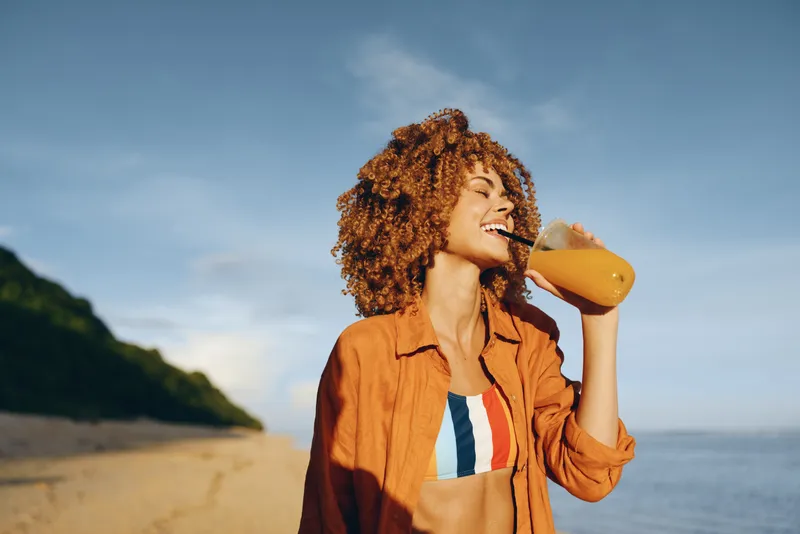woman drinking juice beach outdoors summer sunlight portrait curly jacket, smiling woman on seaside vacation, drinks orange beverage from glass with straw, relaxed healthy lifestyle near ocean, blue