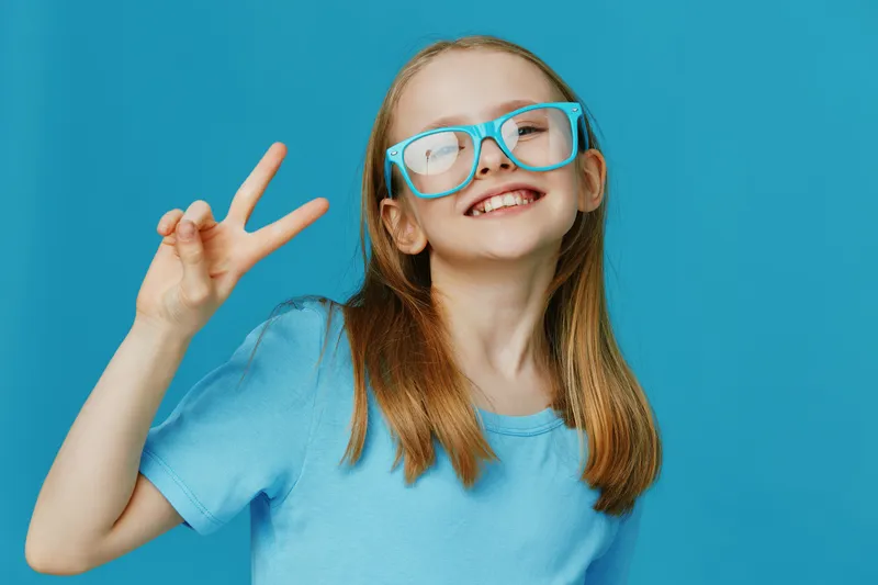Cheerful girl with long hair wearing blue glasses and a blue t shirt, expressing joy with a peace sign on a vibrant blue background