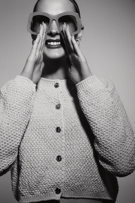 Smiling young white woman with light skin wearing sunglasses and knitted sweater, posing joyfully in a studio. Fashion and happiness concept.