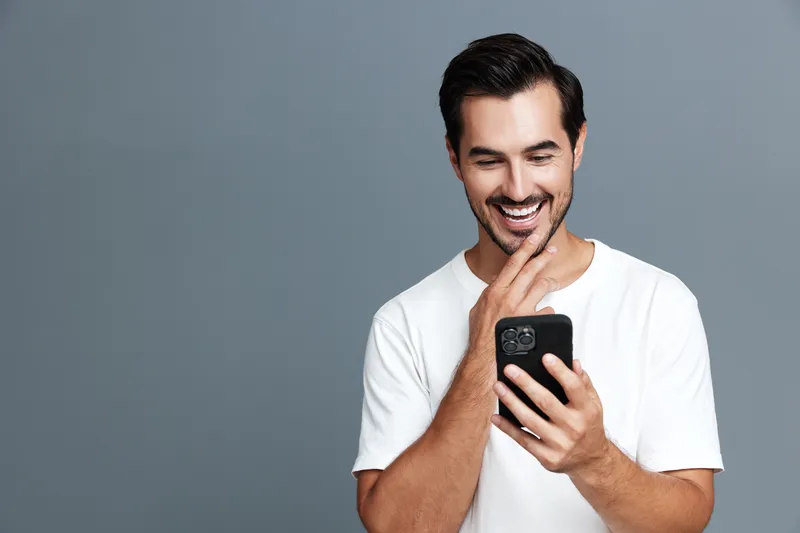 Smiling man in casual white t-shirt looking at smartphone with joy and curiosity, isolated on solid grey background. Modern lifestyle concept.