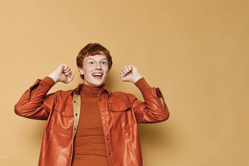 Young man with red curly hair wears orange shirt and jacket smiling with raised fists on beige background. Joyful expression, casual style and positive emotion.