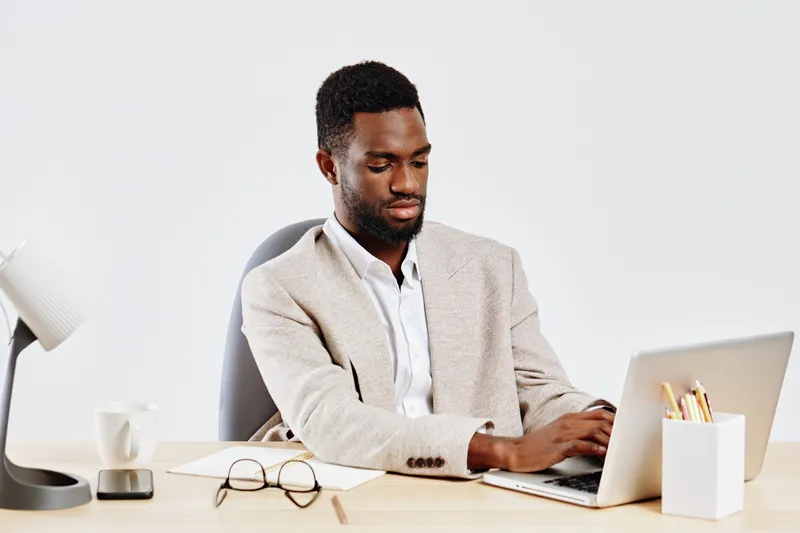 Professional young black man working on a laptop in a modern office, dressed in smart casual, focused expression. Office workspace with accessories, neutral background, business and technology concept