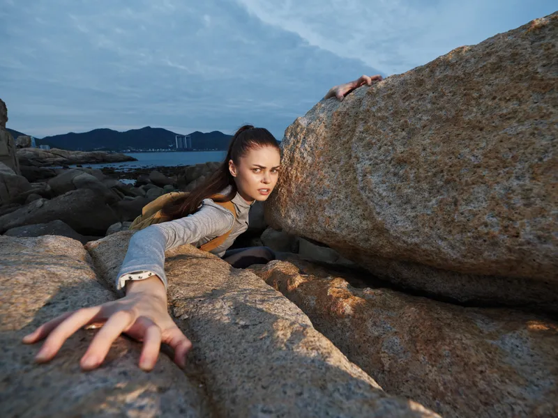 A determined young woman struggling against large rocks by the sea, showcasing emotions of resilience and strength in a dramatic outdoor setting