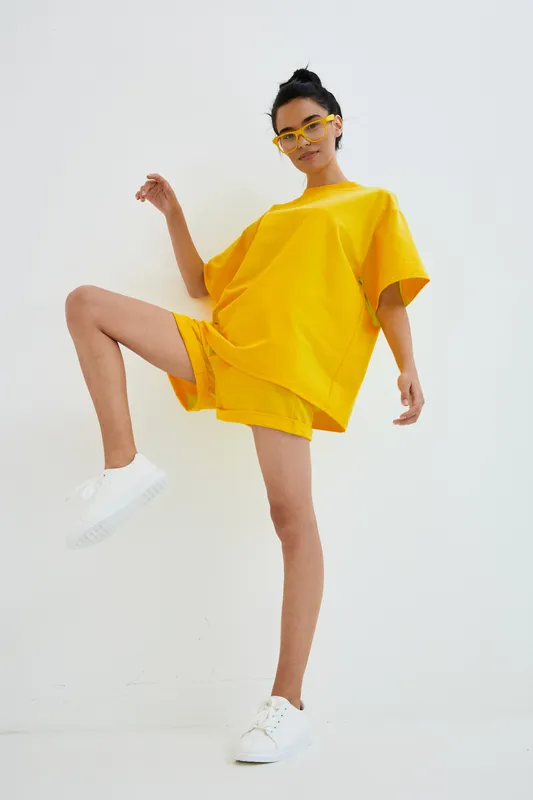 Young woman wearing bright yellow casual outfit and matching sunglasses poses confidently against plain white background, showing energetic summer style and modern streetwear fashion.