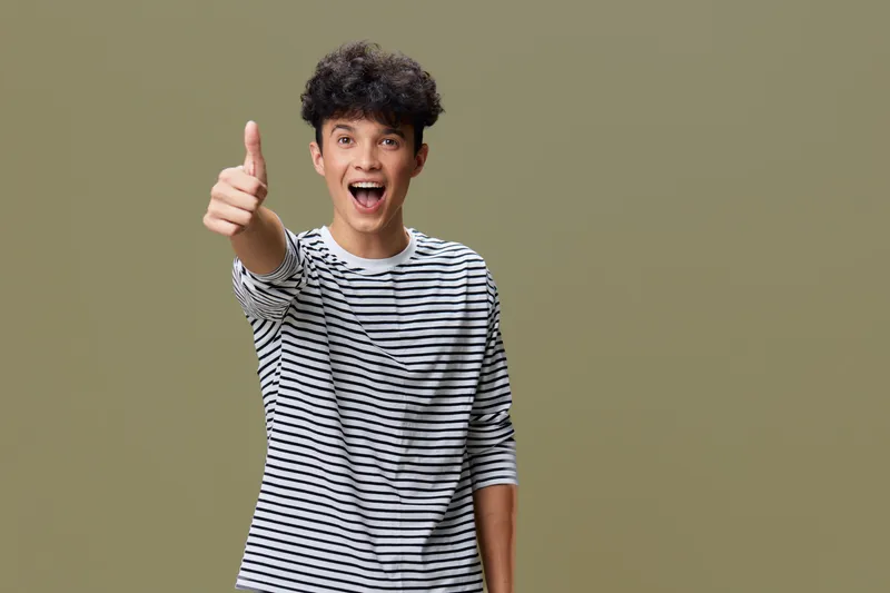 young man showing thumbs up with excited expression wearing striped shirt on beige background, positive gesture and casual style portrait