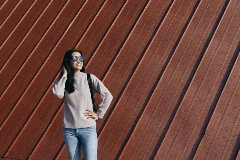 Woman with backpack posing against a bold diagonal wall, wearing sweater and jeans, sunglasses, urban backdrop, casual travel vibe, confident stance, street fashion composition, lifestyle and