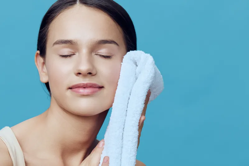 Young woman gently patting her face with a towel, conveying relaxation and skincare, on a calming blue background, with a focus on fresh and healthy skin