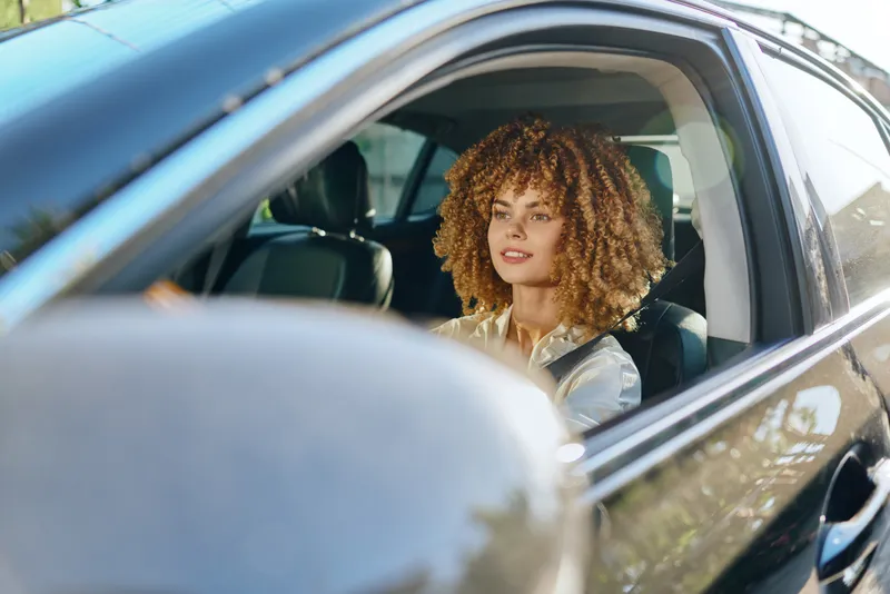 young woman with curly hair driving car, sunlight, casual clothing, focused expression, urban background, daytime scene, modern lifestyle